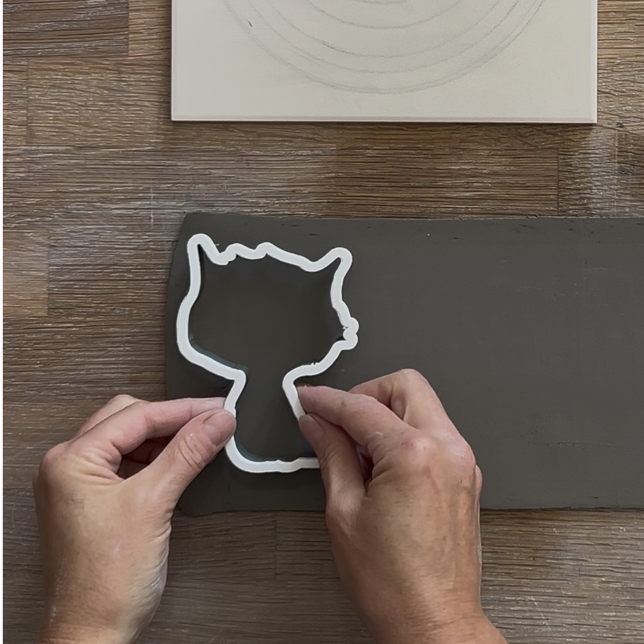 Weißer Halloween-Ausstecher in Form einer schrägen Katze wird auf eine ausgerollte Tonplatte gedrückt, um eine Keramik-Silhouette auszuschneiden.